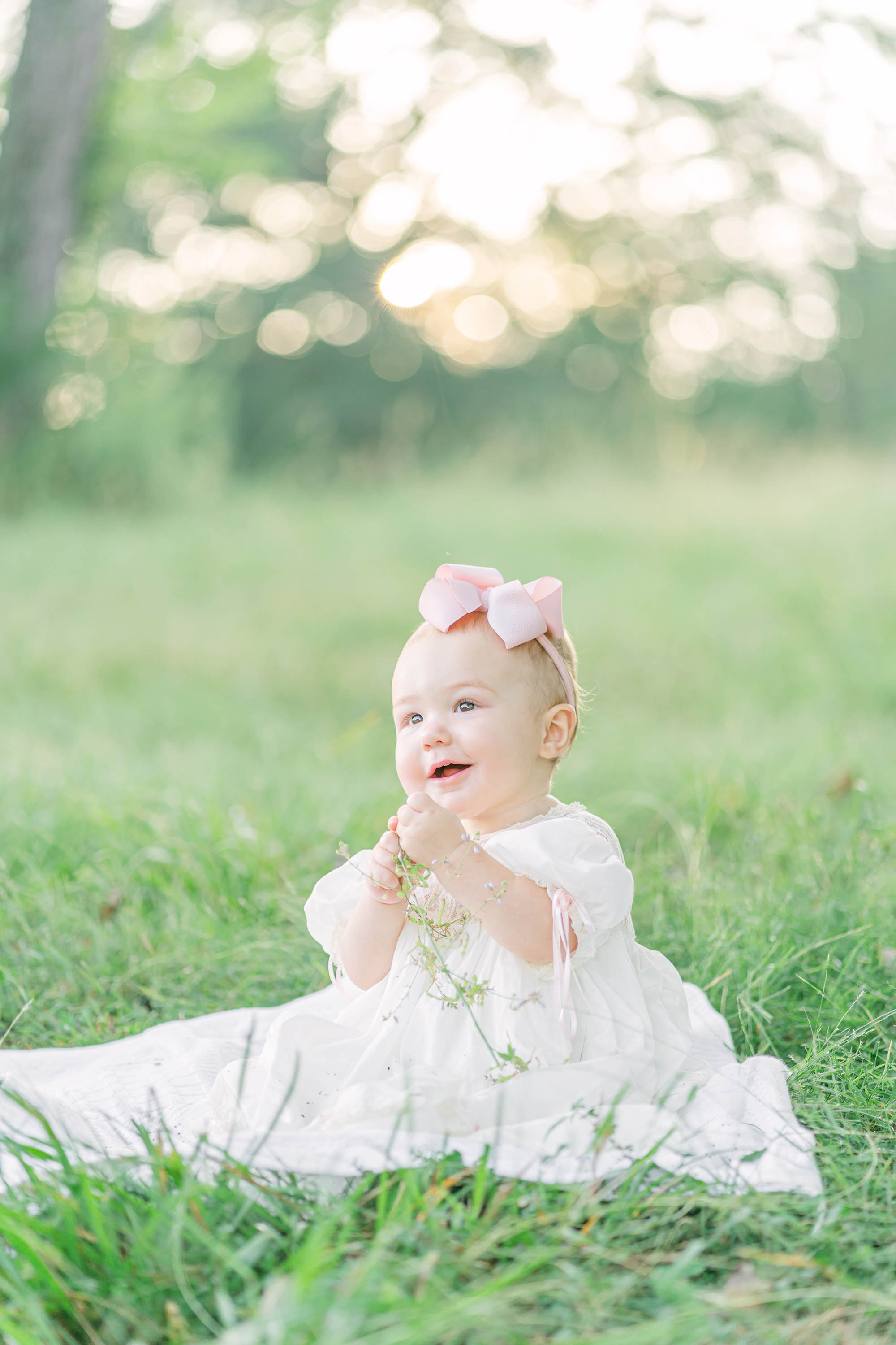 1 year old girl wearing a long white dress sitting in green grass and smiling happily