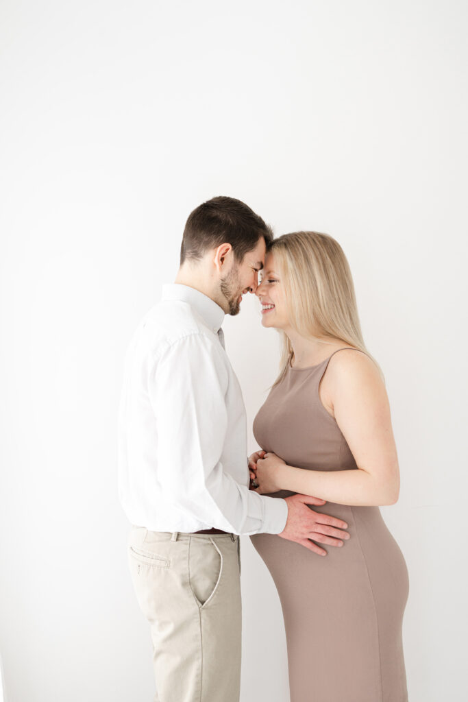 Pregnant mother  with husband 
posing for maternity portraits in a white room wearing a form fitting tan dress.