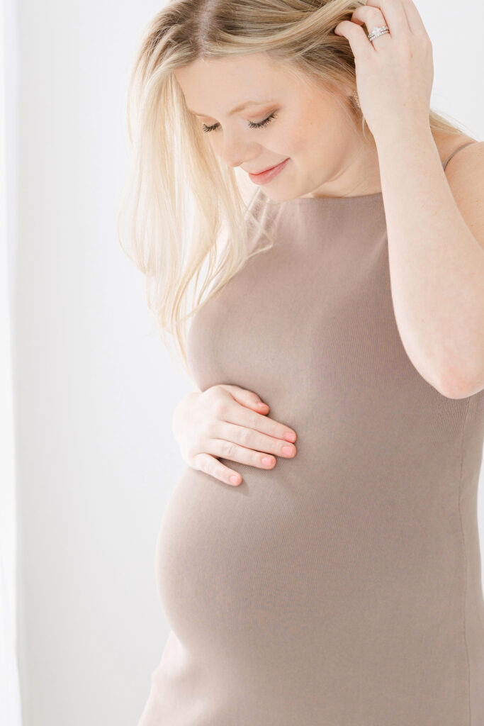 Pregnant mother posing for maternity portraits in a white room wearing a form fitting tan dress.
