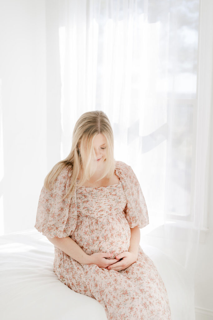 Pregnant mother sitting on a white bed surrounded by beautiful light.  