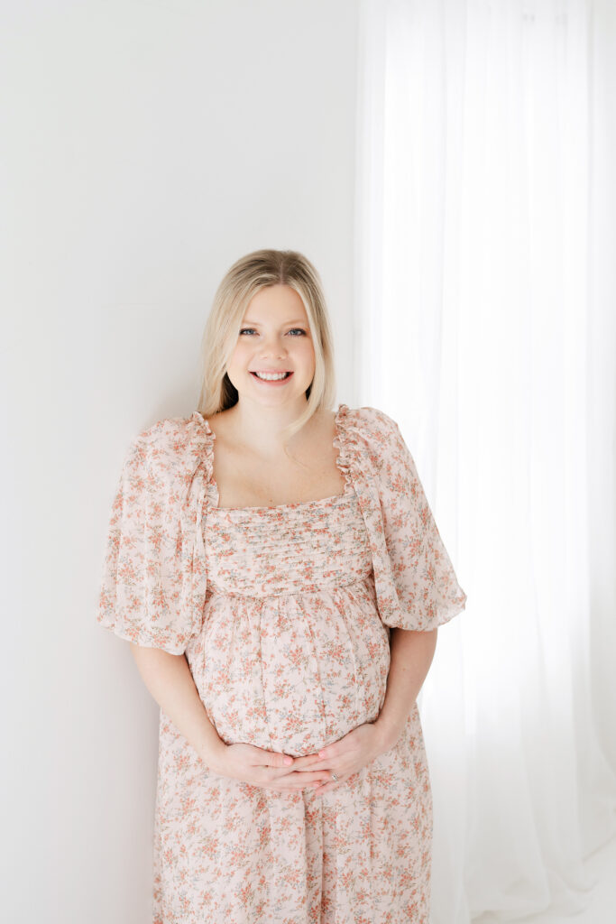 Pregnant mother posing for maternity portraits in a white room wearing a pink dress.