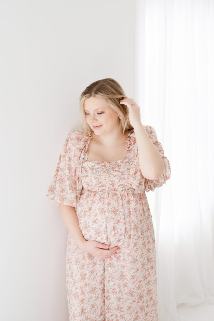 Pregnant mother posing for maternity portraits in a white room wearing a pink dress.