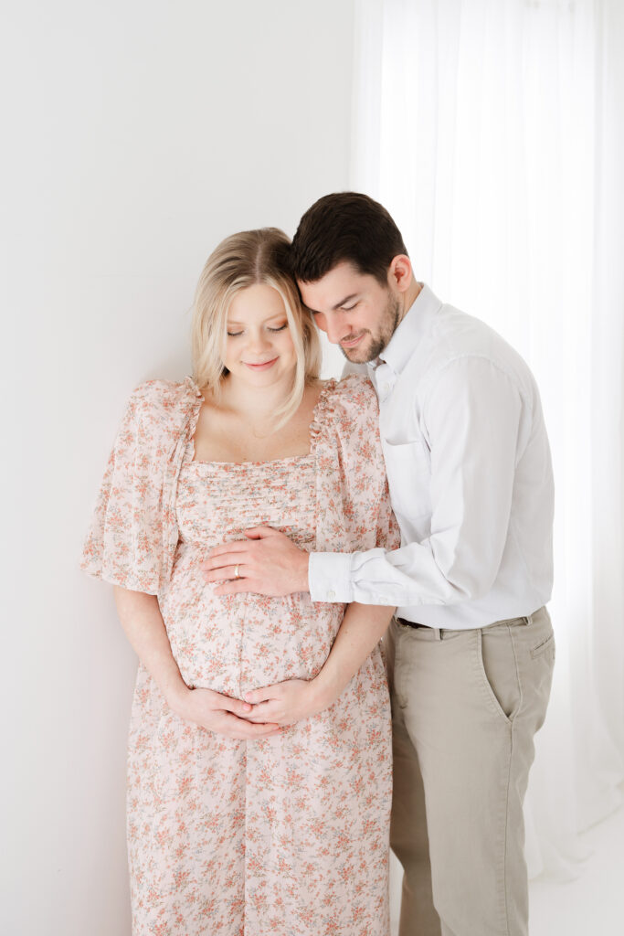 Pregnant mother with husband posing for maternity portraits in a white room wearing a pink dress.