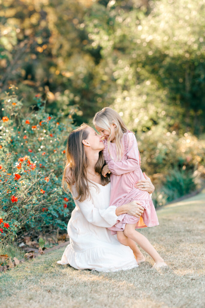 A mom sitting on the ground rubbing noses for a cute picture with her daughter in a green garden.