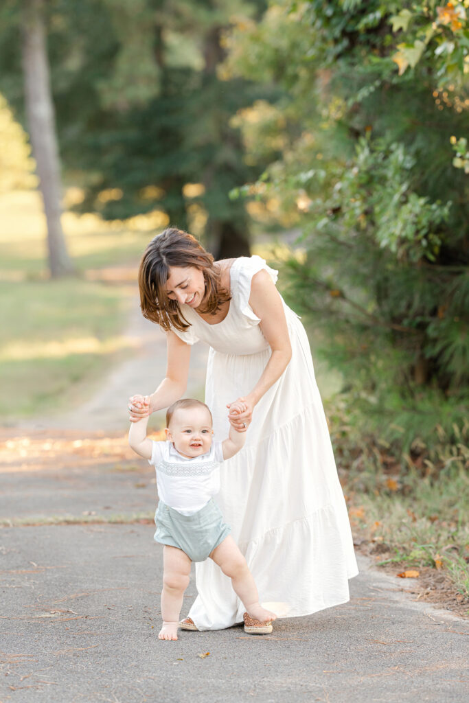 A young mom in a white dress helping her 1 year old, son walk.