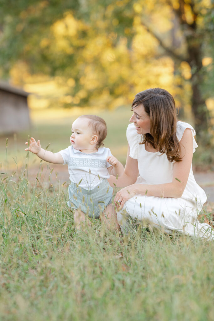 A mom kneeling with her 1 year old baby in a green field while he is amazed with the tall grass.