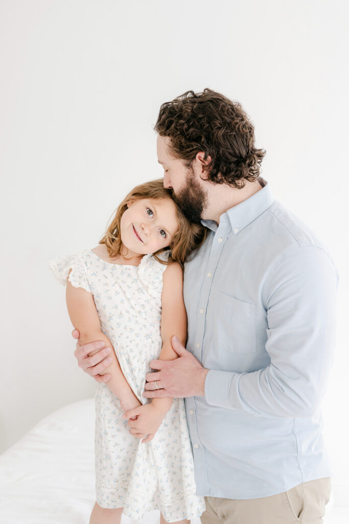 A father kissing his young daughter on the head while posing for a picture in a white room.