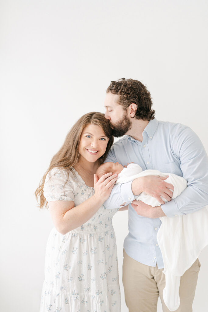 A father kissing his wife on the head while holding his newborn and posing for a picture in a white room.