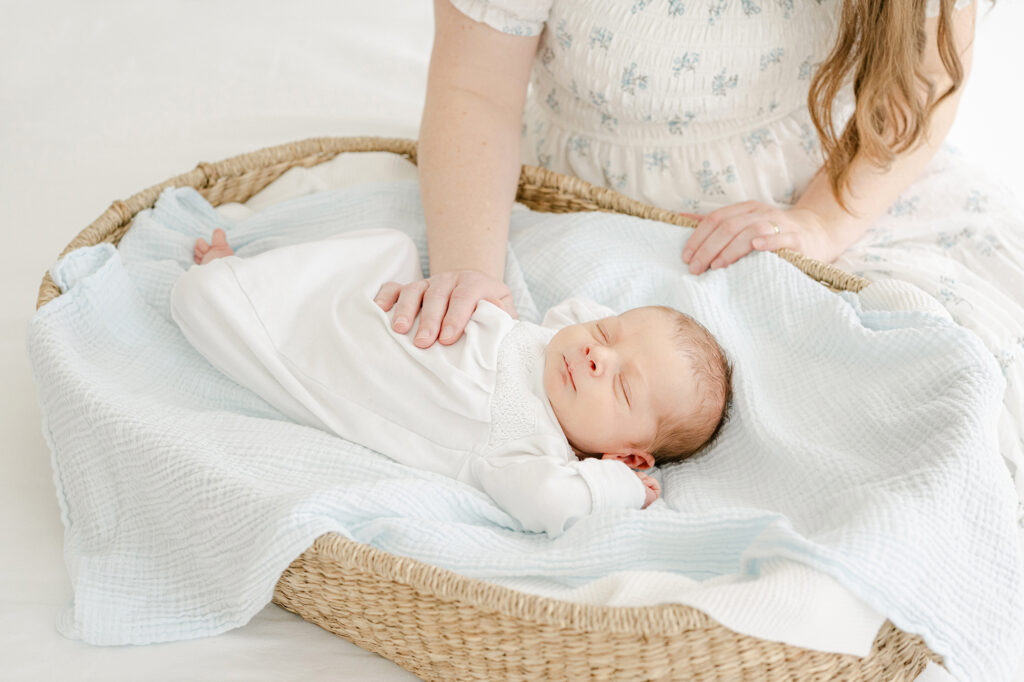 a newborn baby sleeping in a basket while mom places her hand on him for comfort.