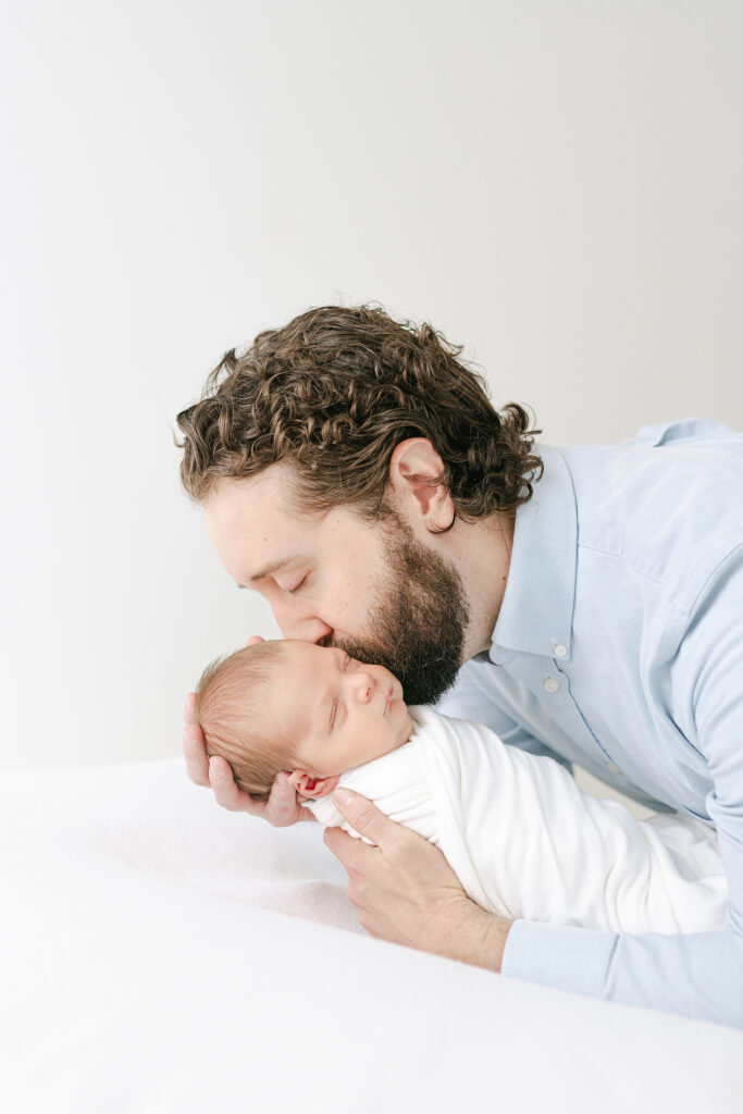 A father kissing his newborn baby on the cheek while posing for a timeless newborn picture in a white room.