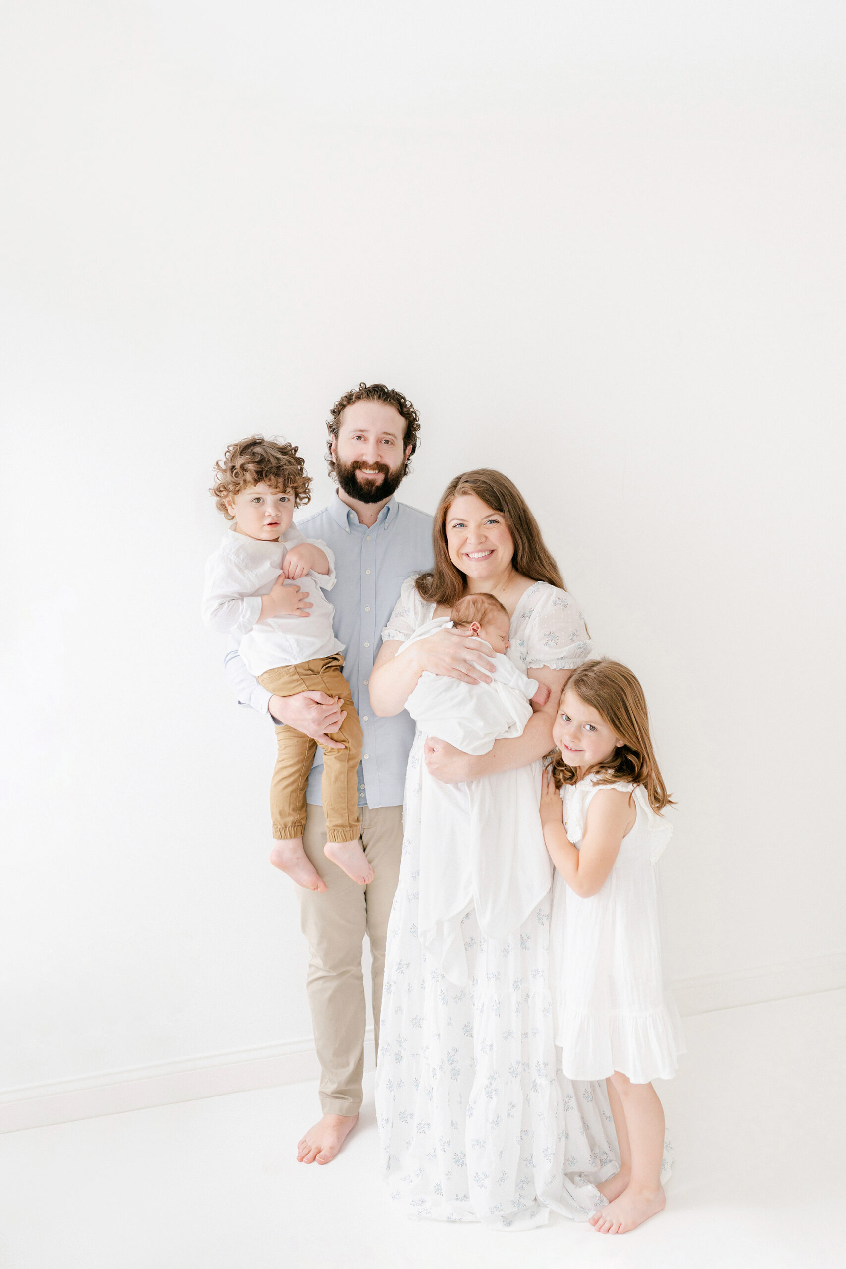 a young family of 5 posing for their fist family photo in a white room with their newest baby.
