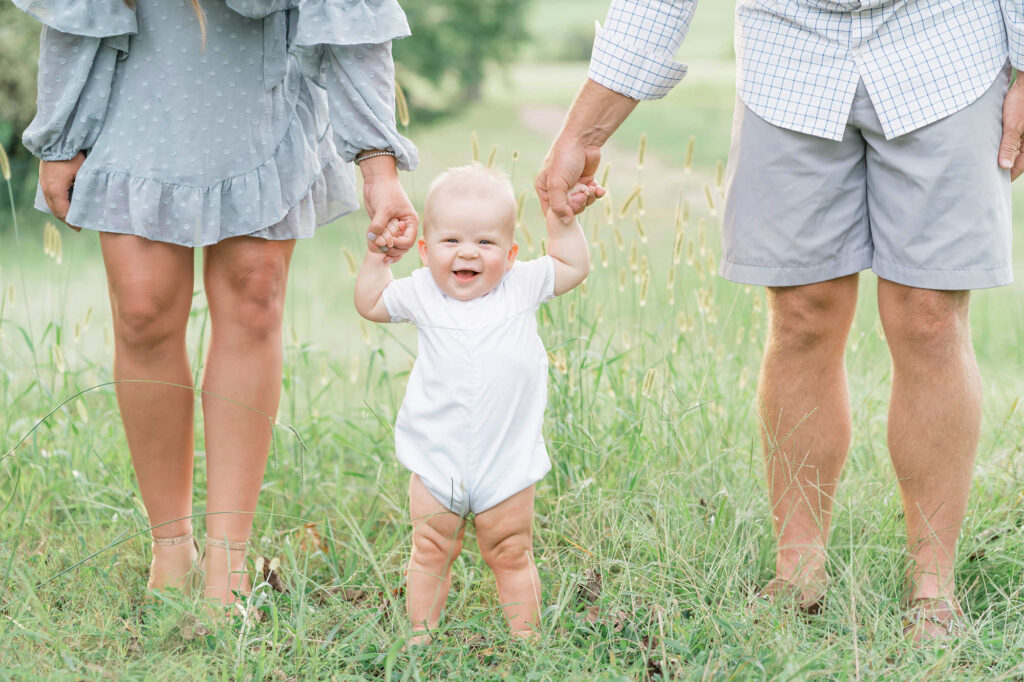 A baby standing in the grass holding both parents hands for support while smiling.