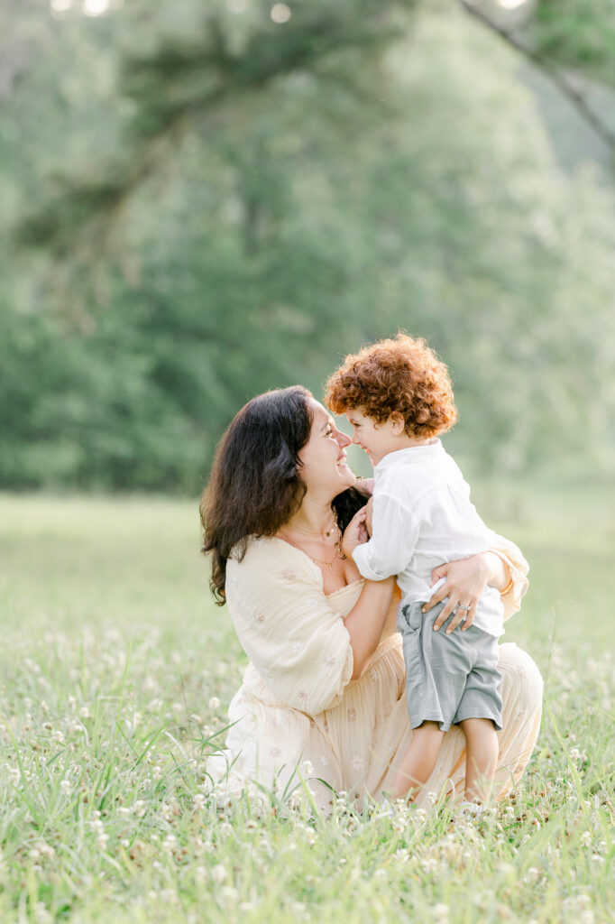 A mom sitting on the ground rubbing noses for a cute picture with her son in a green field.
