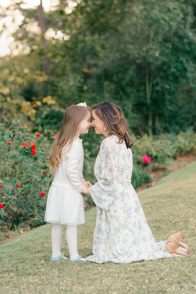A mom kneeling on the ground rubbing noses for a cute picture with her daughter in a green garden.