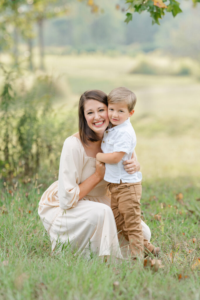 young mom cheek to cheek for a picture with her toddler son in a green field.