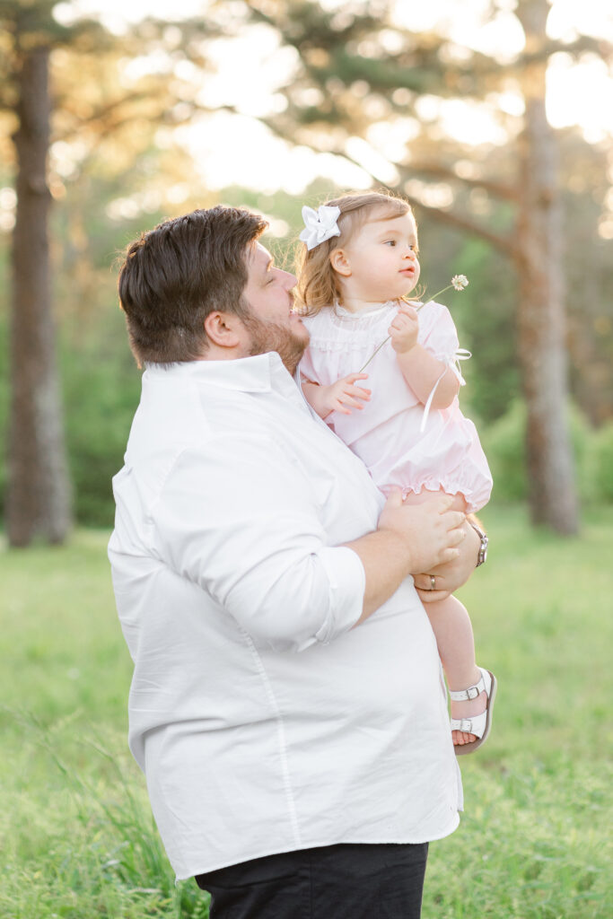 a dad holding his young daughter in a green field while she holds a flower and they look off in the distanc.