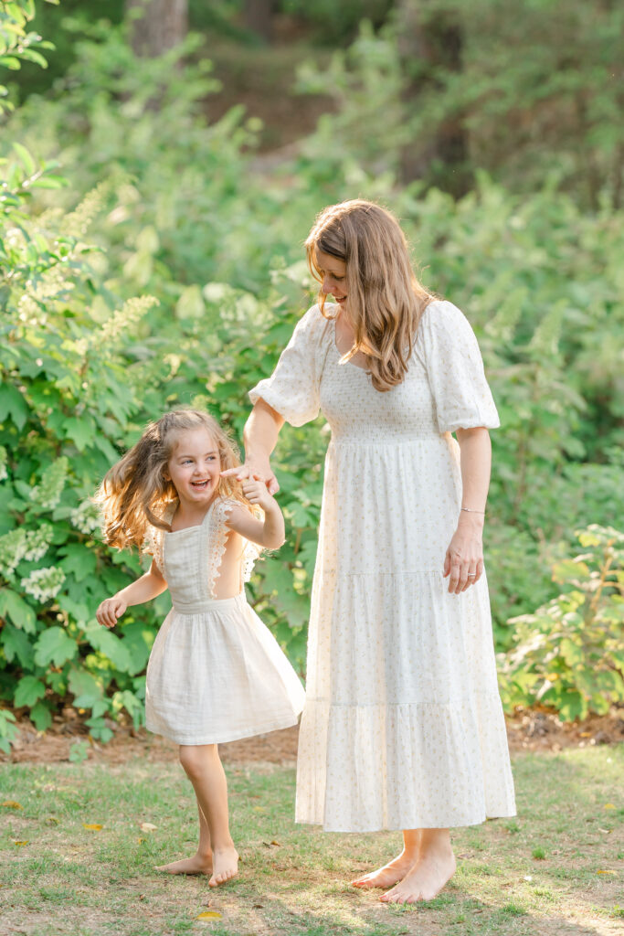 A mom in a white dress twirling her laughing daughter in a green garden.
