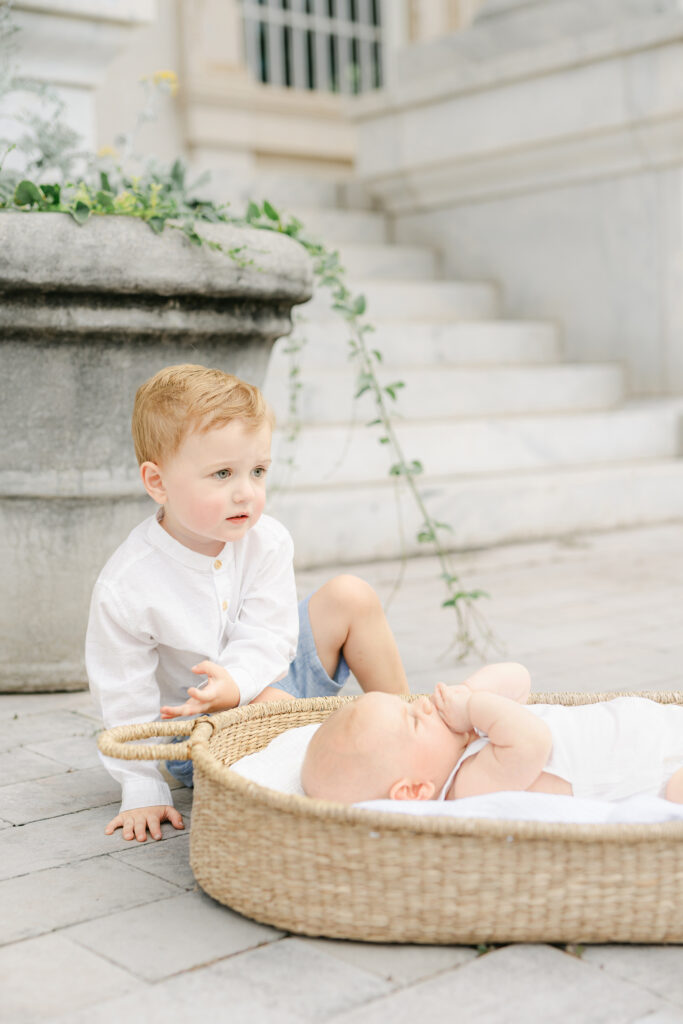 a toddler playing next to his baby brother who is in a basket
