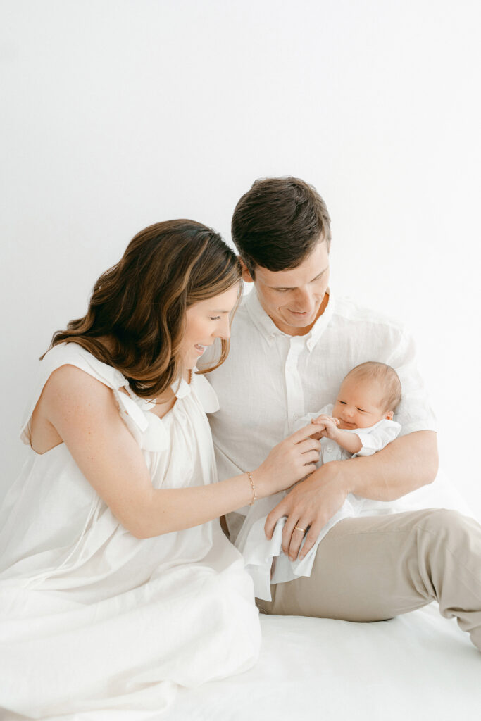 A new mom and dad hold their newborn baby and talk to him while sitting on a bed in a white room.