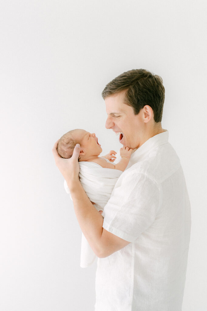 A new dad makes the same  cute face as his newborn son during a photo session in a white room.