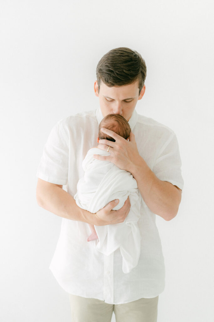 A new dad kisses his newborn on the forehead during a photo session in a white room.  We only see the back of the newborns head.