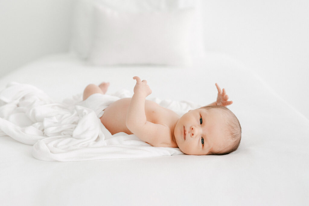 Photo of a 2 week old baby in a white diaper cover looking at the camera while laying on a bed.  