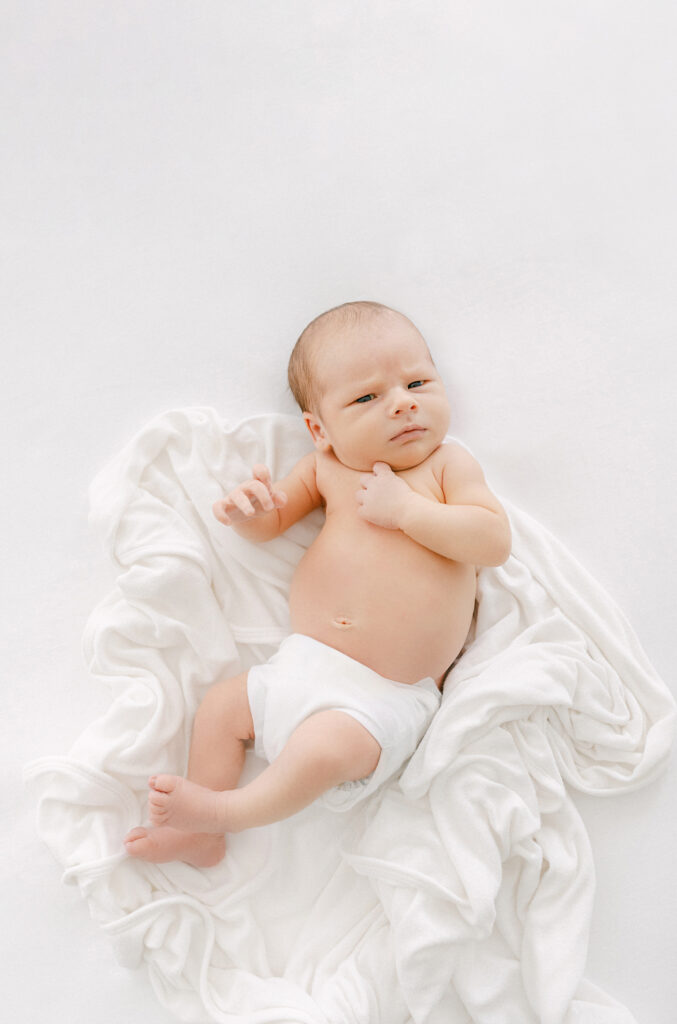 Photo of a 2 week old baby in a white diaper cover looking at the camera while laying on a bed. Photo is taken from above.