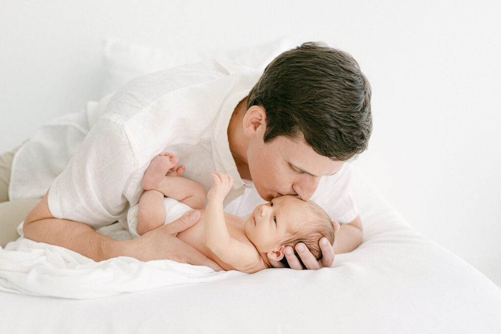 A new dad kisses his newborn on the forehead during a photo session in a white room.  They are both laying on a bed.