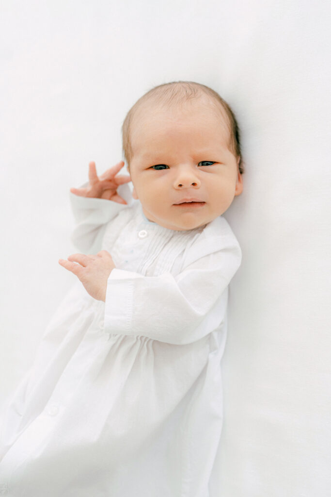 A newborn, dressed in a white, heirloom gown looks at the camera for a portrait.