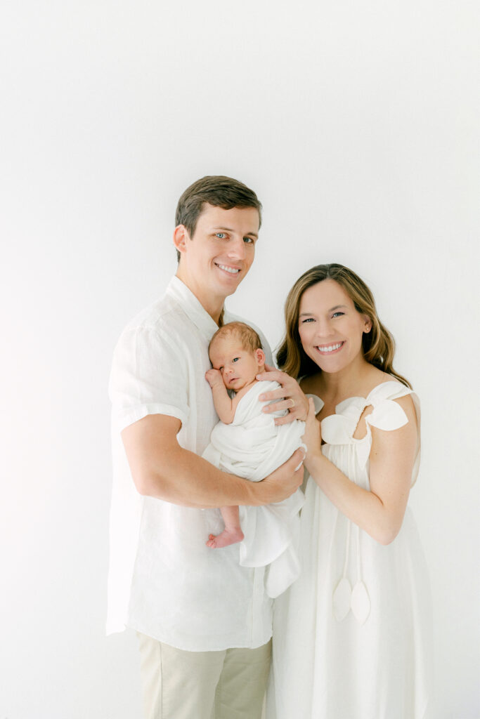 A new mom and dad hold their new baby and pose for a portrait in a white room during a photosession.