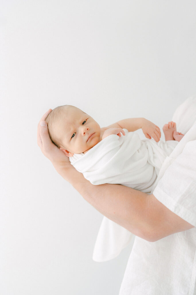 A newborn baby, wrapped in a white blanket looks at the camera for a portrait while propped on dad's chest.