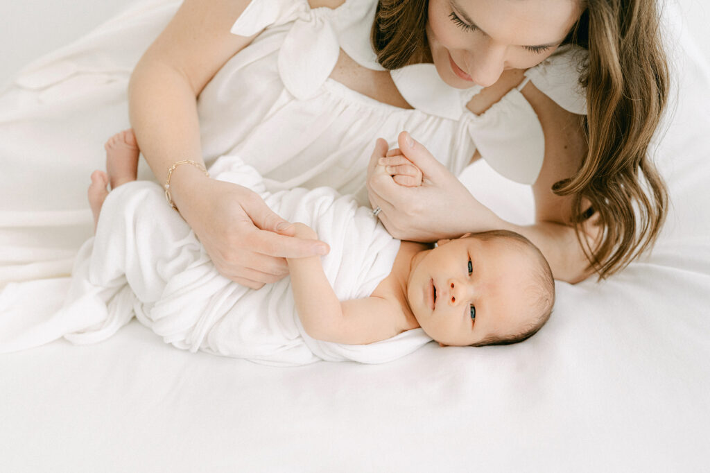 A new mom snuggles her newborn on a bed during a photo session in a white room.