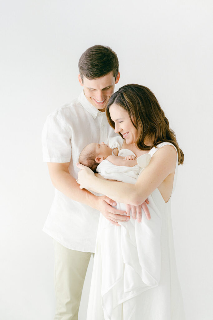 A new mom and dad snuggle their newborn during a photo session in a white room.