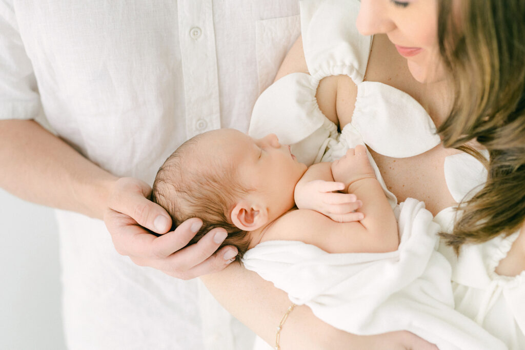 photo of a new mom holding her newborn baby in a white room.  Dad cradles the newborns head in his hand.