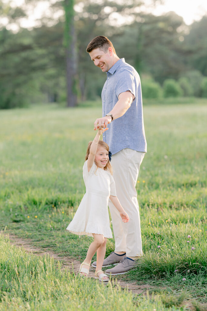 a father twirls his young daughter during an outdoor photo session in a sunlit field.