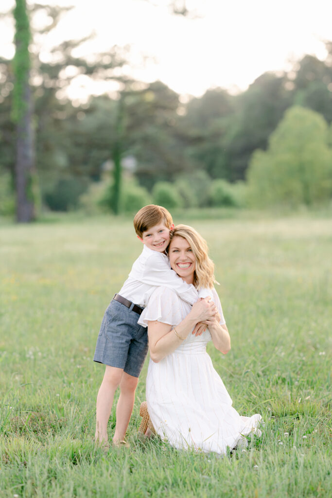 a young son wraps his arms around his mothers neck during a photo session in a sunlit field