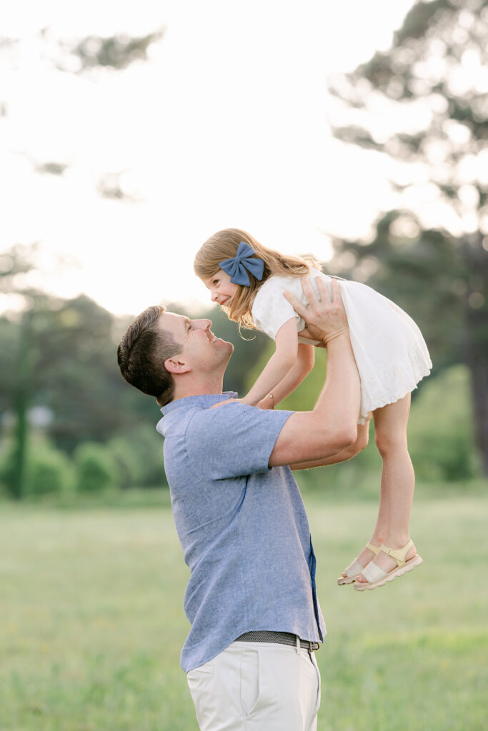 a father holds his young daughter  in the air during an outdoor photo session in a sunlit field.