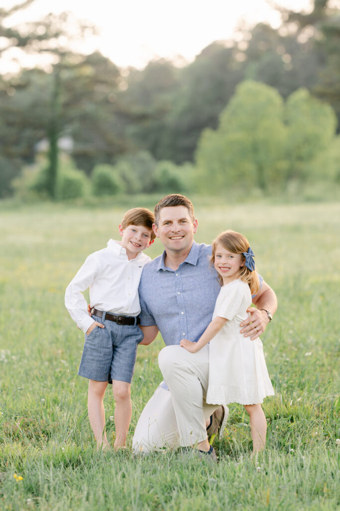 a father hugs her young daughter and son during a photo session in a sunlit field