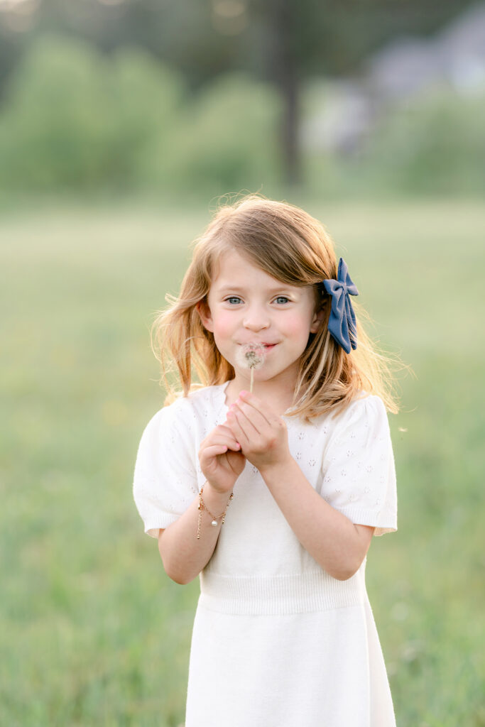 a young girls blows a dandelion in a field while smiling for the camera
