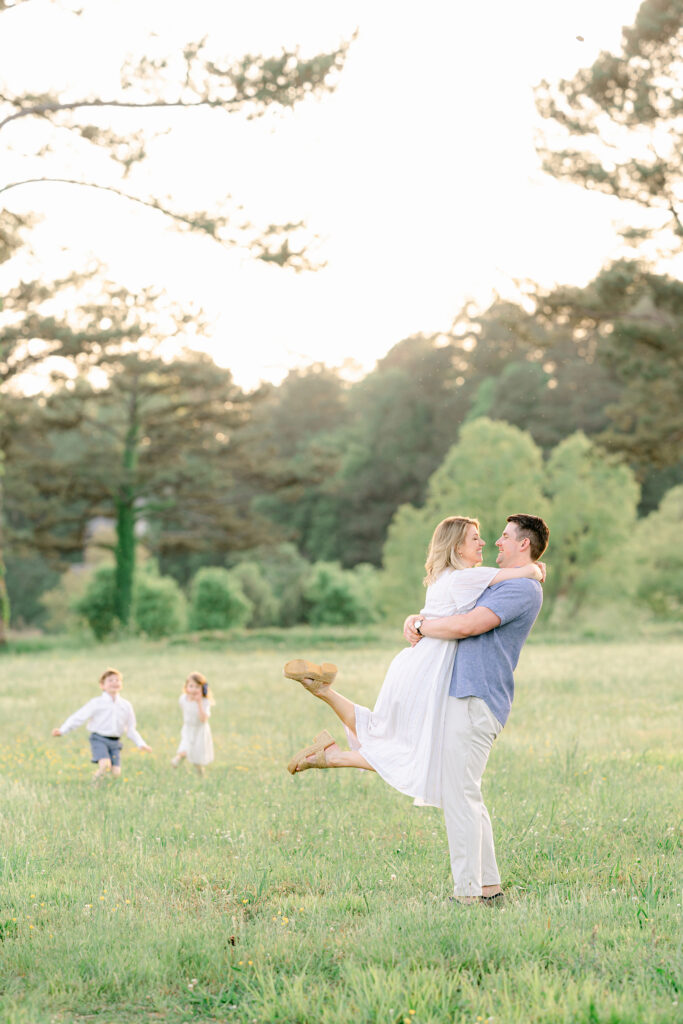 a mom and dad embrace in a fun way while their young children play in the background
