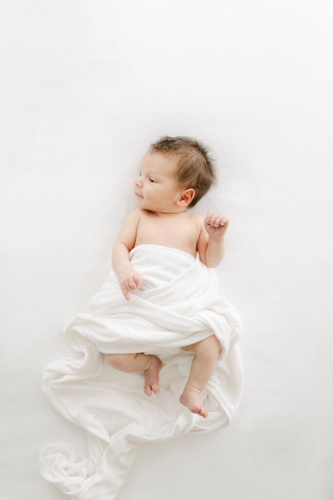 a new baby relaxes and smiles on a white  bed during his photosession.