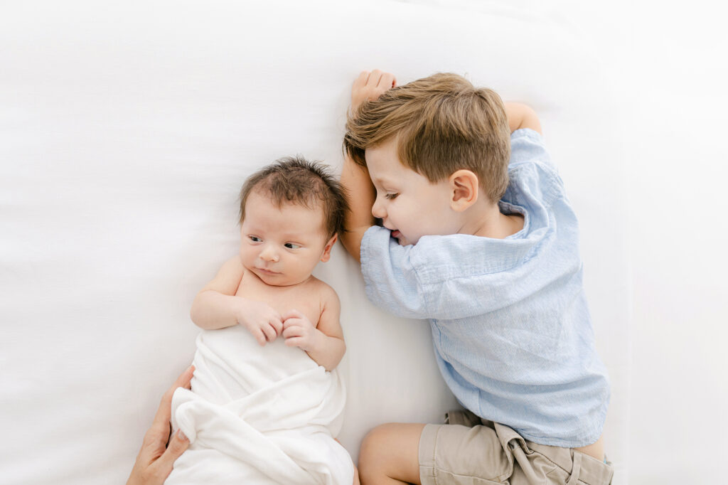 a new baby relaxes on a white  bed during his photo session while his big brother smiles at him