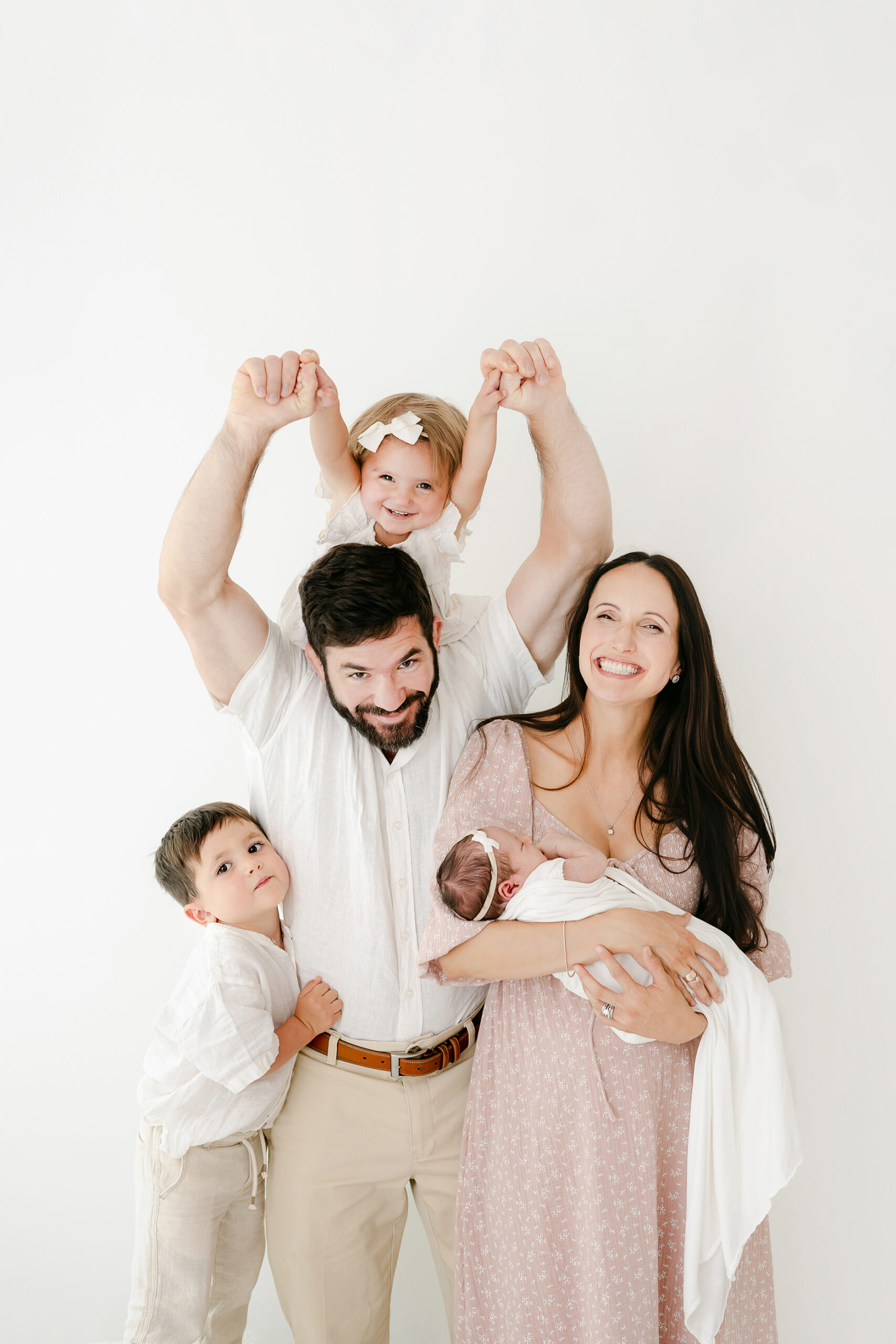 family of 5 with 2 toddlers and a newborn. One toddler is on Dad's shoulders whie the other hugs him and mom smiles while holding the newborn.